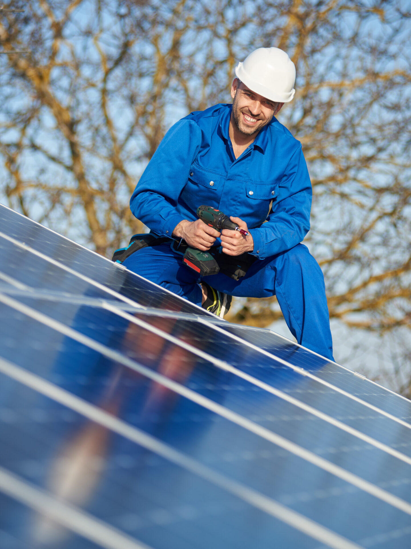 Electrician mounting solar panel on roof of modern house Portrait of man worker in blue suit and protective helmet installing stand-alone solar photovoltaic panel system using screwdriver. Smiling electrician mounting blue solar module on roof of house.